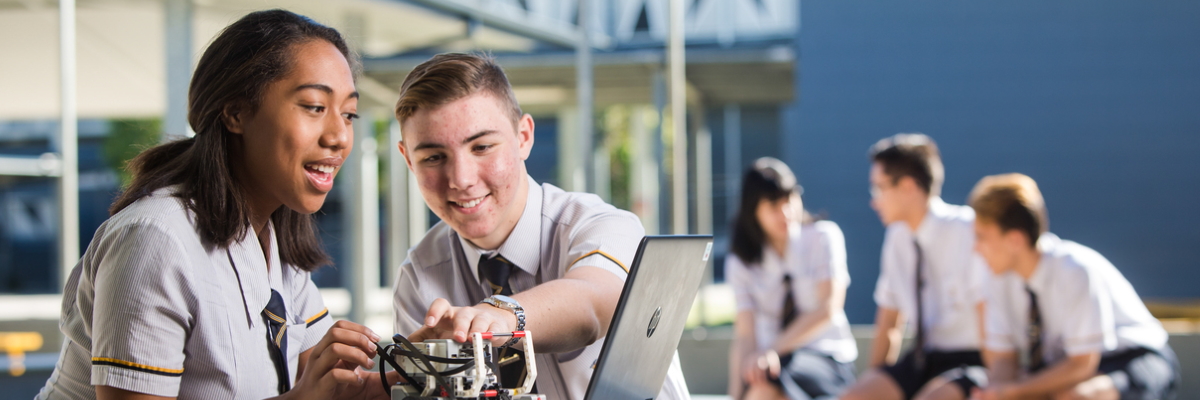 Two students doing robotics with a group of more students in the background
